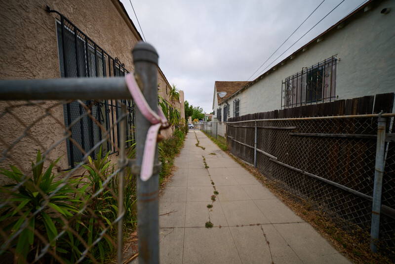 Narrow residential alleyway with chain-link fences, brick buildings, and overcast sky.