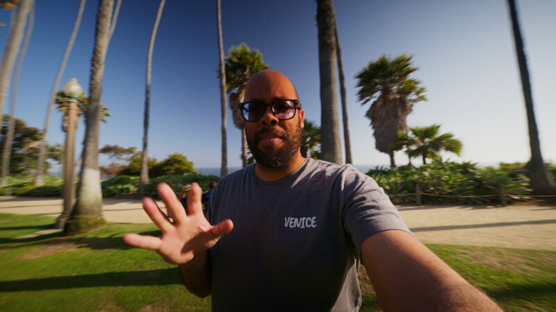 Man wearing glasses and Venice t-shirt gesturing toward camera in palm tree-lined park during daytime.