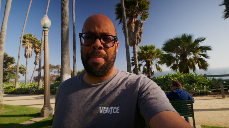 Man wearing sunglasses and Venice-branded t-shirt standing outdoors with palm trees behind him.