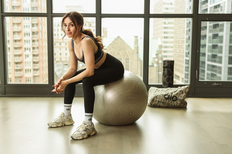 Woman in black athletic wear seated on a stability ball in a high-rise gym with floor-to-ceiling windows overlooking a city skyline.
