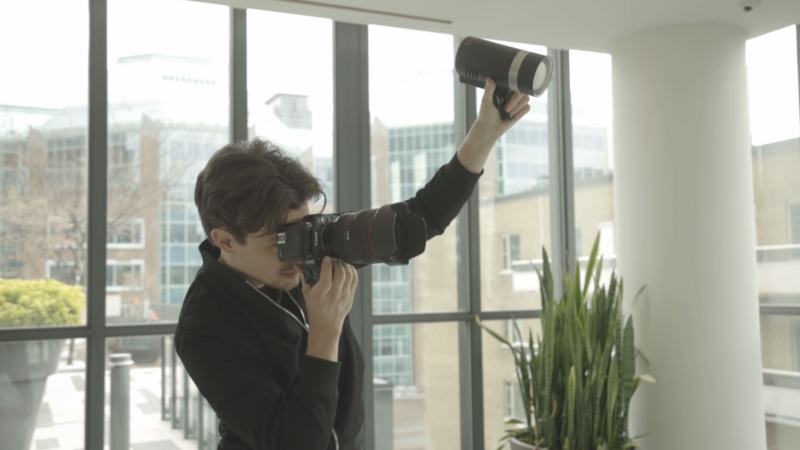 Photographer holding camera overhead to capture a high-angle shot through large windows overlooking a cityscape.