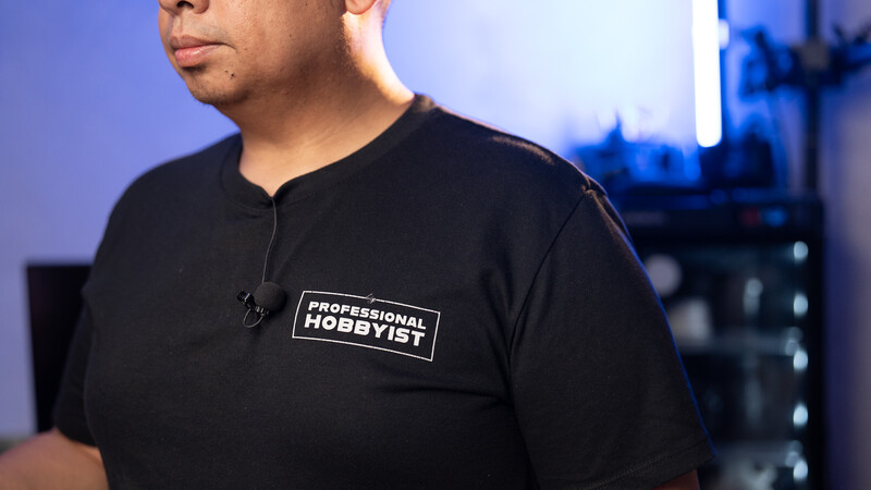 Man wearing black shirt with "Professional Hobbyist" text patch, photographed indoors with city lights visible in background.