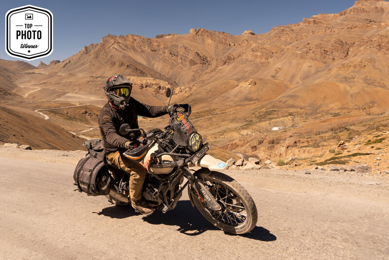Motorcycle rider in helmet and jacket poses on vintage bike in arid desert landscape.
