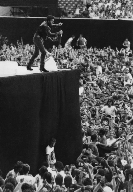 Performer on elevated stage platform with large crowd visible below in black and white photograph.