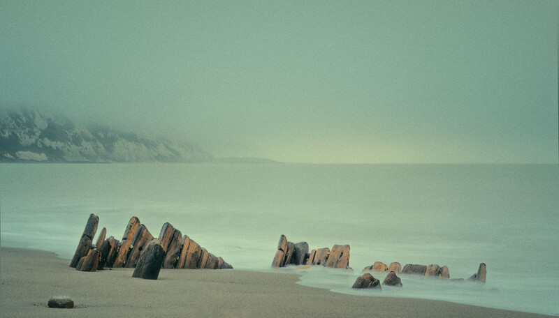 Weathered wooden shipwreck remains partially submerged on a misty beach.