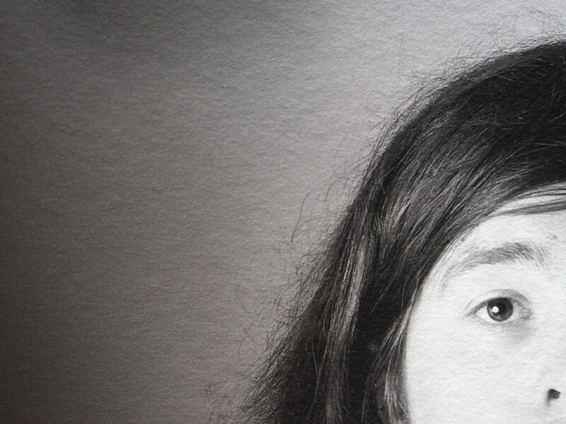 Close-up black and white portrait showing partial face with dark hair and one visible eye against neutral background.