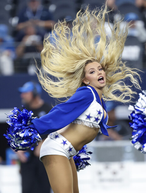 Cheerleader in mid-jump wearing blue and white uniform with blonde hair in motion.