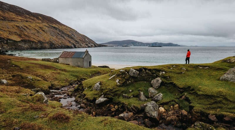 Solitary hiker in red jacket stands on grassy coastline overlooking calm bay with small cottage and mountains beyond.