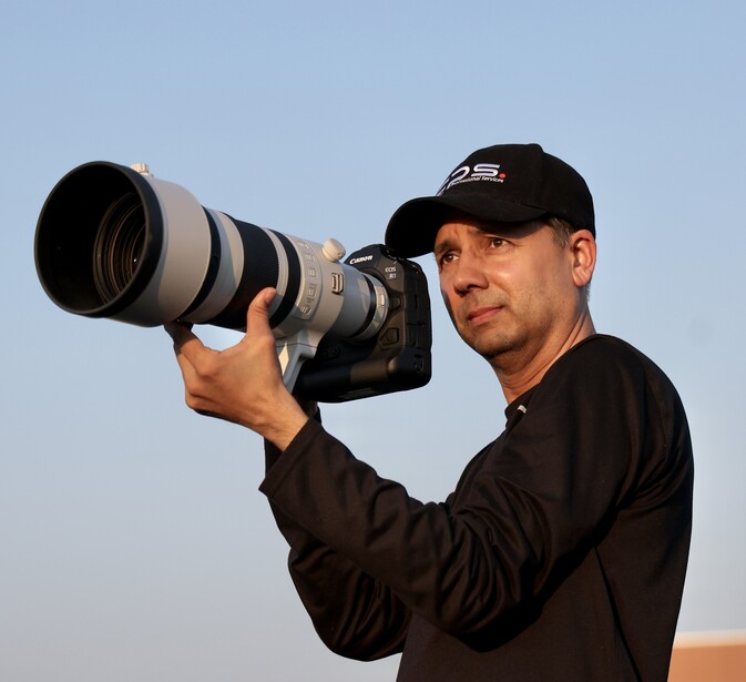 Male photographer holding a telephoto lens camera against clear sky background.