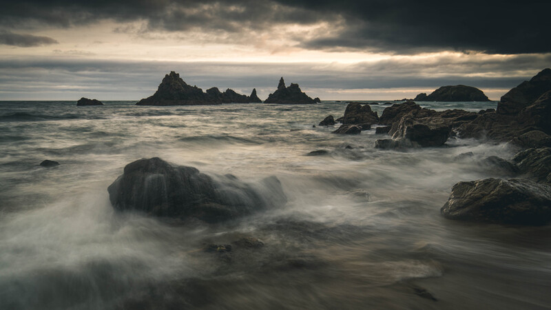 Rocky coastline with silhouetted sea stacks and waves washing over dark boulders under dramatic cloudy sky.