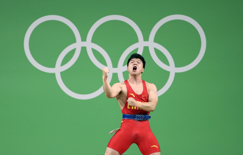 Weightlifter in red competition attire celebrating with fist raised against Olympic rings backdrop.