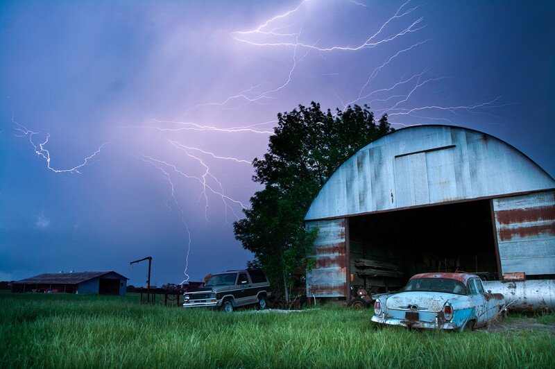 Lightning streaks across a stormy sky above a weathered metal barn and abandoned vintage car in a rural field.
