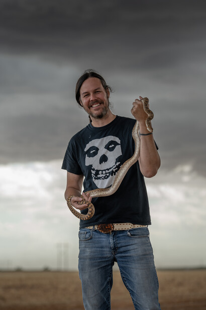 Man in skull graphic t-shirt standing in open field under dramatic cloudy sky.