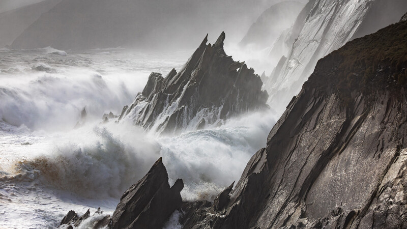 Dramatic rocky coastline with jagged peaks surrounded by crashing waves and sea spray in monochrome.
