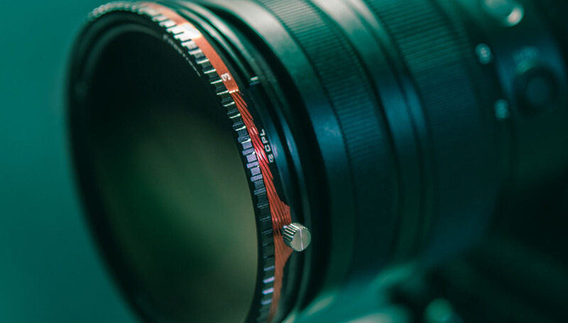 Close-up of camera lens internals showing glass elements and red mechanical components against teal background.