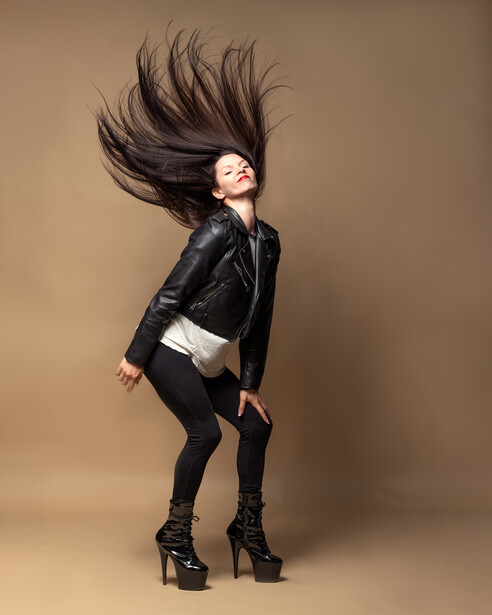 Woman in black leather jacket with long dark hair in motion against tan background.