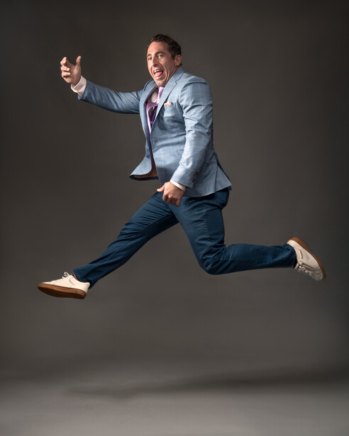 Man in business casual attire jumping with thumbs up gesture against gray background.