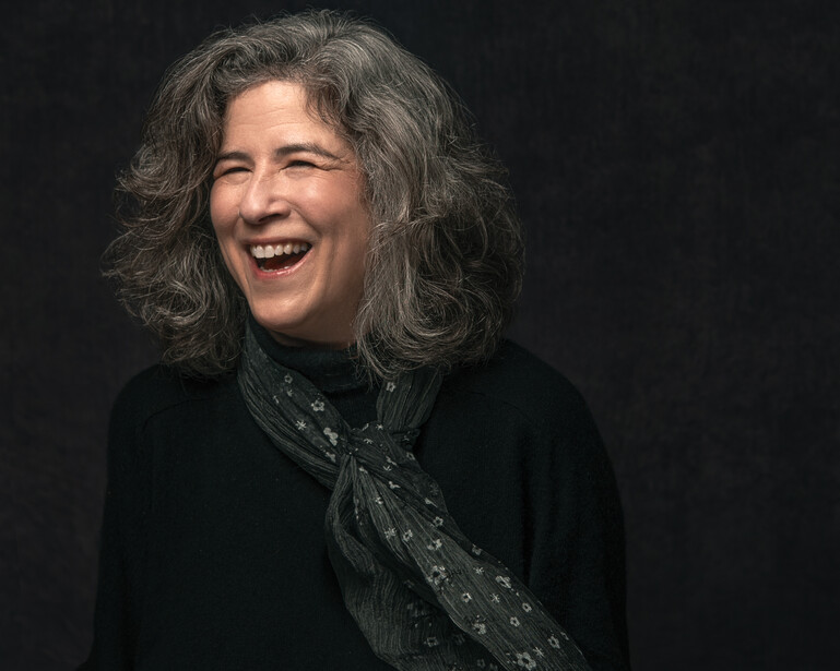 Woman with long gray hair laughing against a dark background.