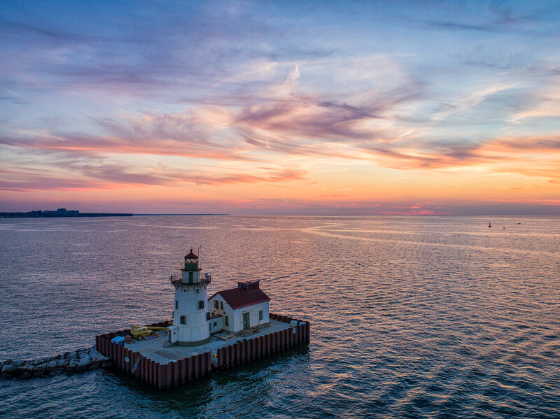 White lighthouse with red roof on wooden pilings at sunset over calm water.