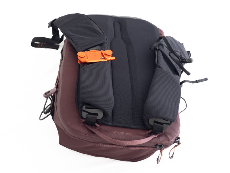 Burgundy and charcoal camera backpack with orange accent straps and side pockets, photographed against a white background.