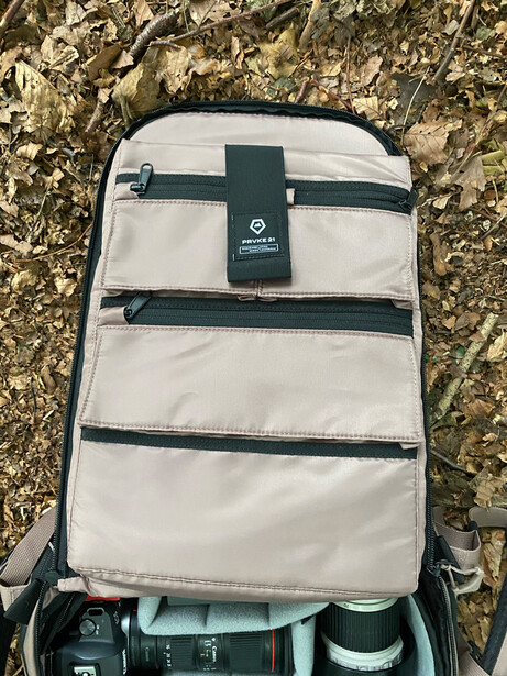 Cream-colored camera backpack with dark straps sitting on textured ground next to camera gear.