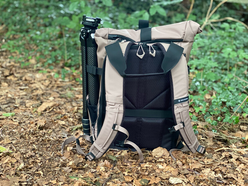 Tan and black roll-top camera backpack displayed from rear view on outdoor ground.