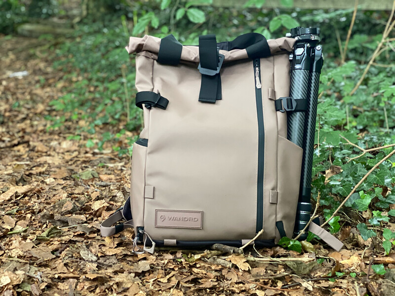 Tan canvas backpack with black straps and hardware displayed on forest floor with green foliage in background.