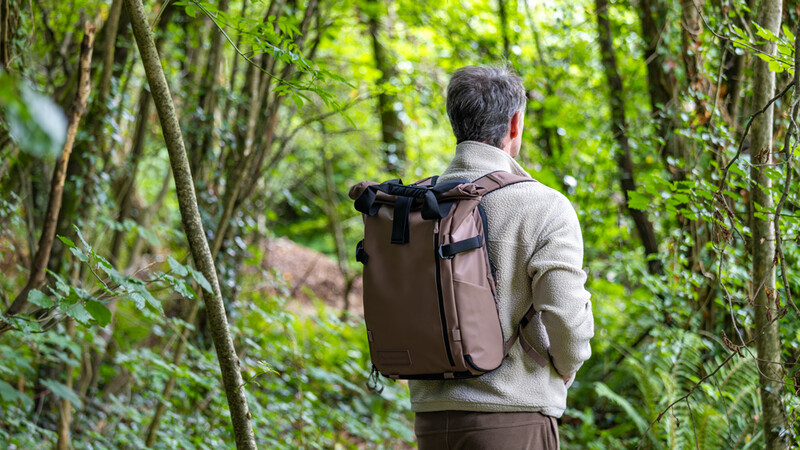 Hiker wearing a khaki backpack walking through a dense forest tunnel of green vegetation.