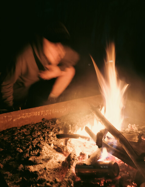 Person leaning over a campfire at night, illuminated by warm firelight against dark surroundings.