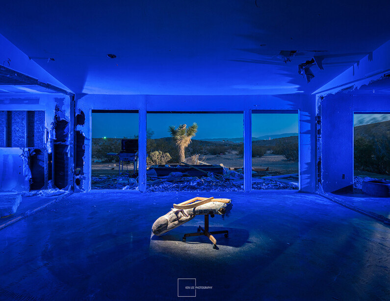 Person in yoga pose on blue-lit floor with expansive windows overlooking mountainous landscape at dusk.