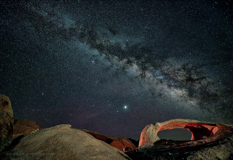 Milky Way galaxy stretching across dark night sky above illuminated rock formations in desert landscape.