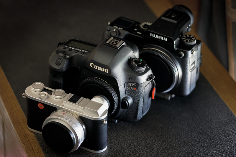 Three cameras arranged on a dark surface: a Canon EOS R5 Mark II, a Fujifilm mirrorless camera, and a compact rangefinder camera.