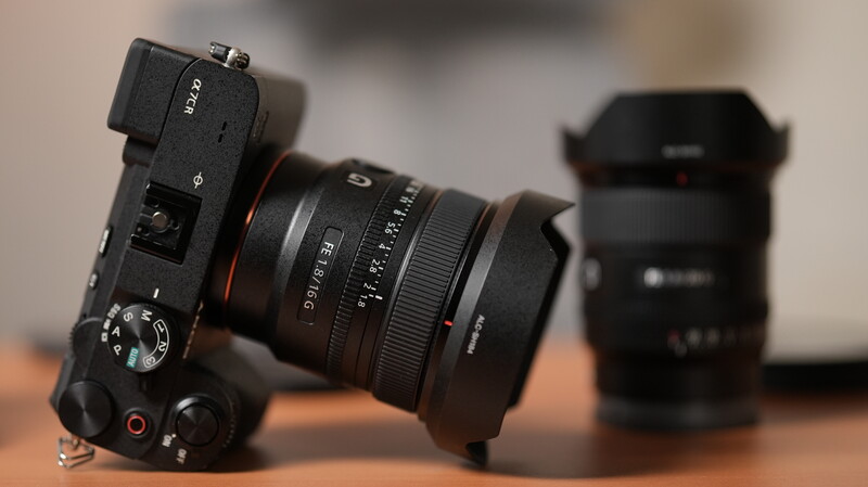 Black mirrorless camera with prime lens mounted, photographed at an angle with additional lens blurred in background.
