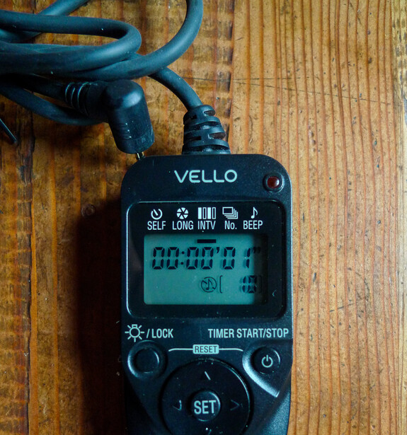 Vello digital light meter displaying exposure readings on a wooden surface with attached blue sync cable.