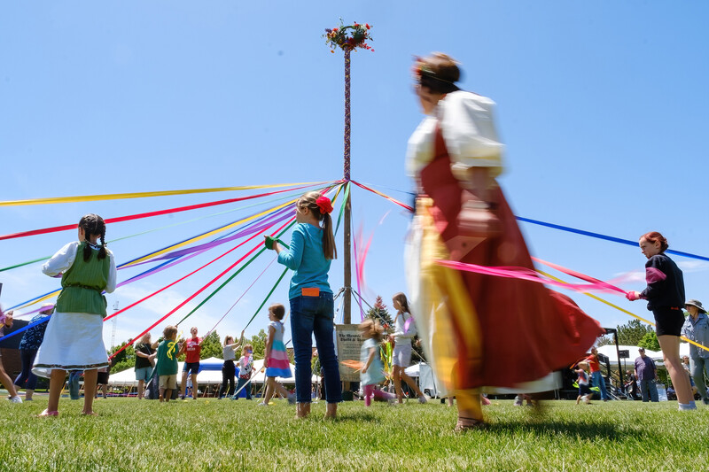 Children and adults participate in a colorful maypole dance outdoors on a sunny day.
