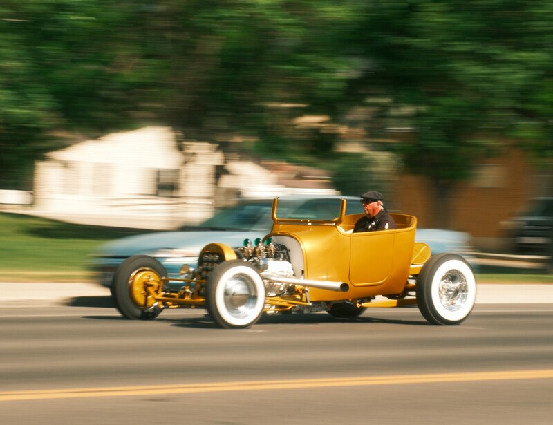 Yellow hot rod with exposed engine driving on road with blurred background.