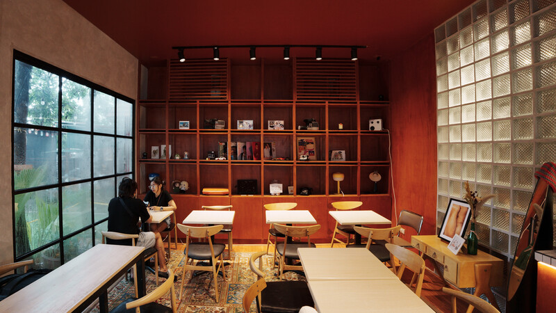 Modern studio interior with warm red walls, wooden shelving, natural light from grid windows, and work tables with chairs.