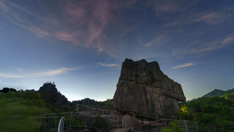 Dramatic rocky pinnacle illuminated at dusk with starry sky above and green hillsides below.