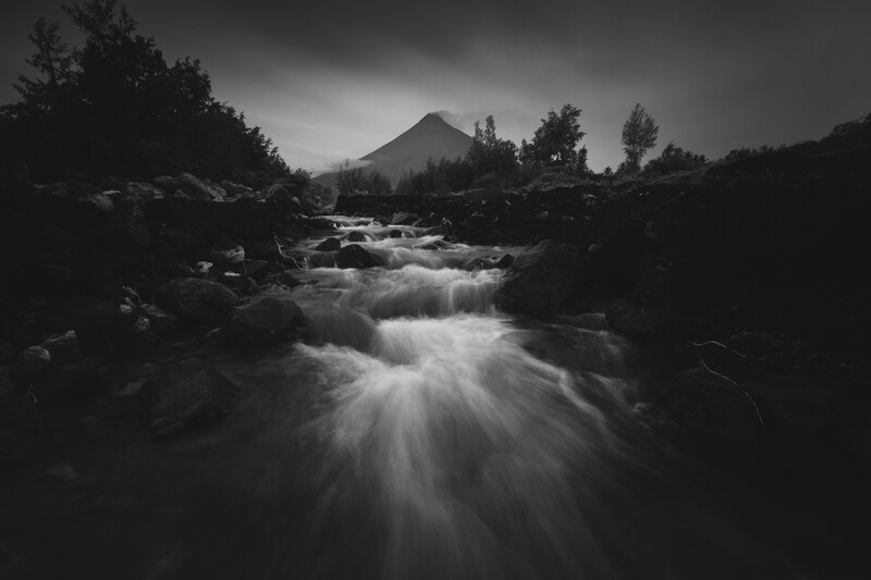 Rushing river with rocks and coniferous forest beneath a volcanic peak shrouded in storm clouds.
