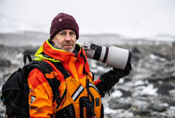 Photographer in bright orange jacket and maroon beanie holding a telephoto lens while standing in a rocky landscape.