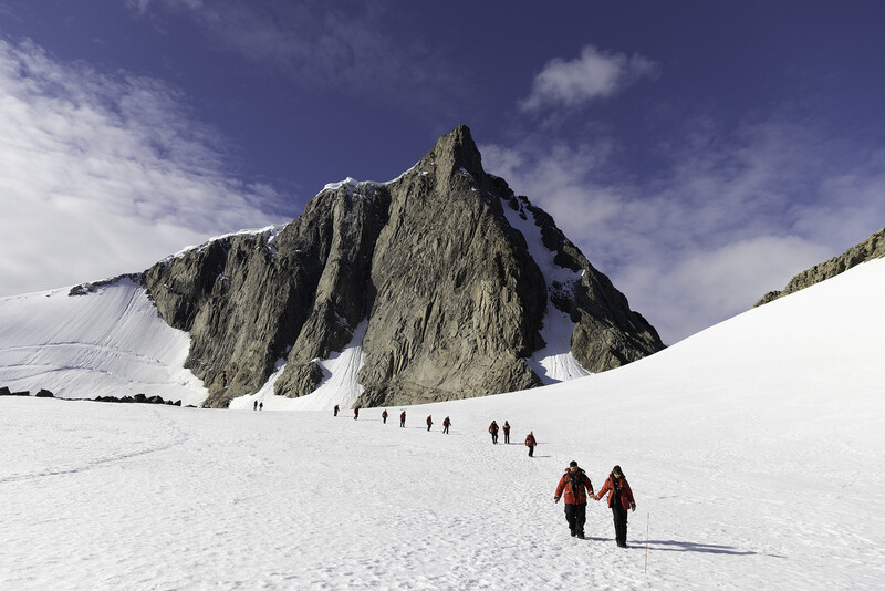 Hikers traversing a vast snowy glacier with a dramatic rocky peak rising in the background.