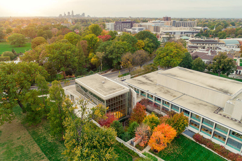 Aerial view of a modern institutional building with flat roofs surrounded by autumn trees and distant city skyline.