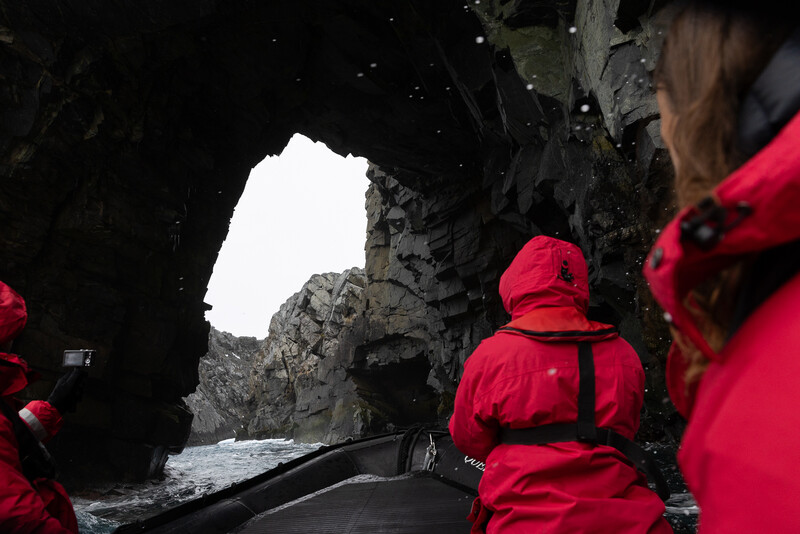 People in red jackets in a boat viewed through a dark cave opening toward bright daylight.