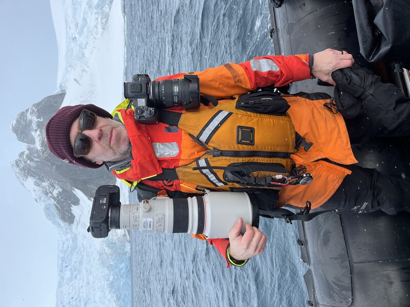 Photographer in orange life jacket holding telephoto lens while seated in inflatable boat with snow-covered mountains behind.