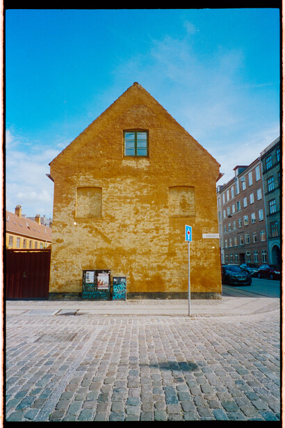 Golden-yellow brick building with symmetrical facade and single window, set in paved plaza against blue sky.