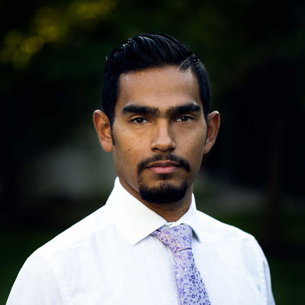 Professional headshot of a man wearing a white dress shirt and patterned tie against a softly blurred green background.