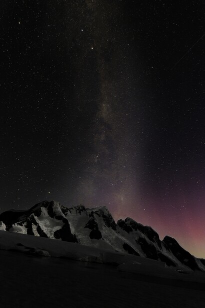 Snow-covered mountain peak silhouetted against starry night sky with Milky Way and purple aurora glow.