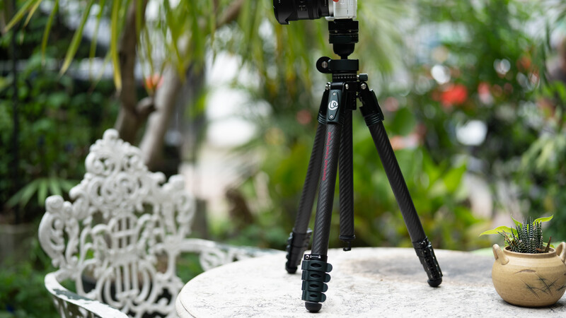 Carbon fiber tripod with ball head mounted on outdoor table surrounded by potted plants and garden decor.
