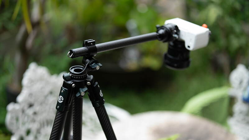 Professional camera boom arm with ball head mounted on carbon fiber tripod legs against blurred garden background.