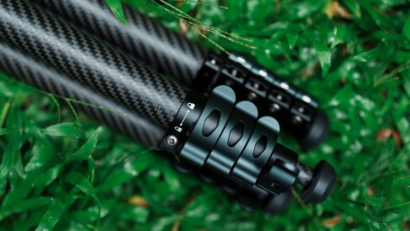 Black carbon fiber camera lens barrel with focus ring on green grass and foliage background.
